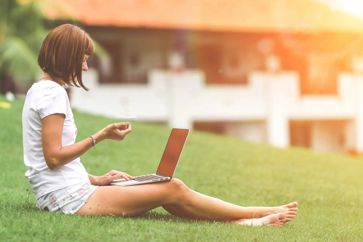 Persoon zit met een laptop op schoot op het gras in de tuin.