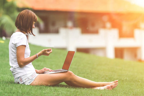 Persoon zit met een laptop op schoot op het gras in de tuin.
