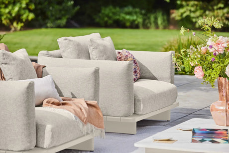 Lichte loungebank en fauteuil met sierkussens op een terras naast het gazon.