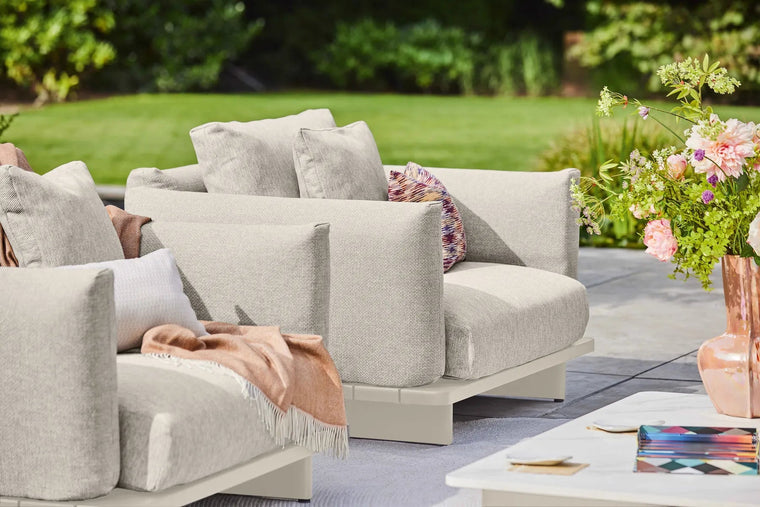 Lichte loungebank en fauteuil met sierkussens op een terras naast het gazon.